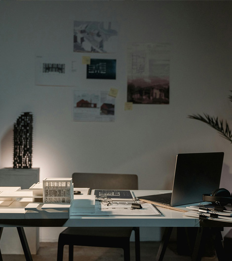 A desk with documents and laptop in a dimly lit room with architectural posters on the wall.